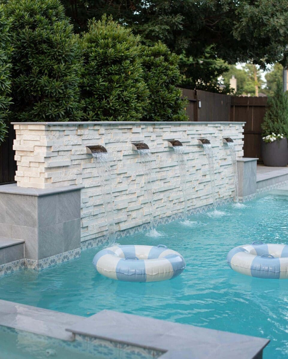 MSI Natural Stone Cosmic White Stacked Stone formed in a wall next to a pool with sheer descent waterfalls. There are tubes floating in the pool.